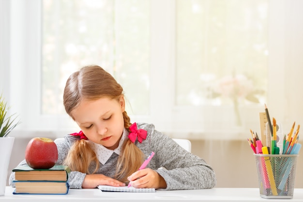 Petite fille étudiante assise à la table et écrit dans un cahier sur un arrière-plan flou.