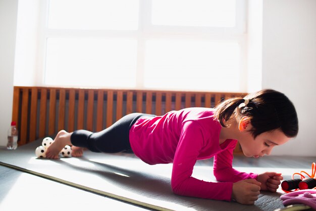 Petite fille faisant une séance d'entraînement Plank Exercise à la maison. Un enfant mignon s'entraîne sur un tapis à l'intérieur. Petite mannequin brune en tenue de sport a des exercices près de la fenêtre dans sa chambre