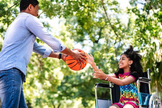Une petite fille en fauteuil roulant s'amusant avec son père tout en jouant au basket-ball ensemble au parc.