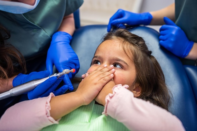 La petite fille ferme sa bouche avec ses mains sur la chaise dentaire du docteur