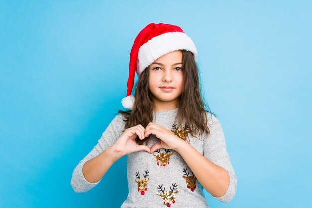 Petite fille fête le jour de Noël en souriant et en montrant une forme de coeur avec les mains.