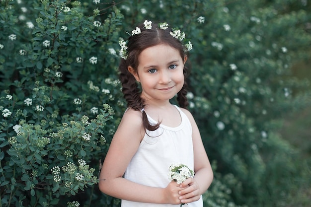 Une petite fille avec des fleurs blanches dans les mains et les cheveux, regarde la caméra et sourit.