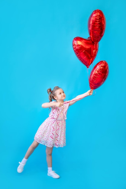 Une petite fille sur un fond de studio isolé bleu avec des ballons rouges en forme de coeur s'amuse et s'amuse Espace pour copier du texte Concept de la Saint-Valentin