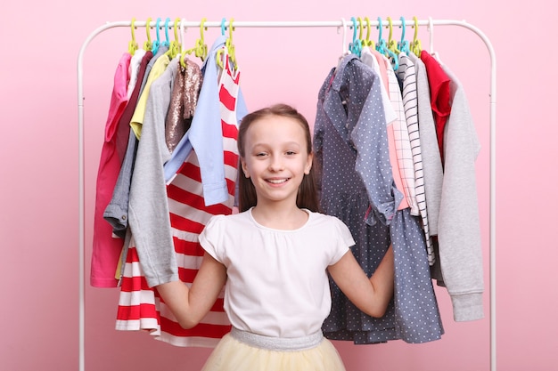 La petite fille gaie mignonne choisit des vêtements avec des cintres de plancher