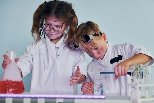Une petite fille et un garçon en blouse blanche jouent un scientifique en laboratoire en utilisant un équipement