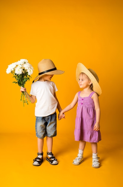 Petite fille et un garçon en chapeaux de paille se tiennent la main sur une surface jaune avec un espace pour le texte. Un garçon tient un bouquet de fleurs blanches