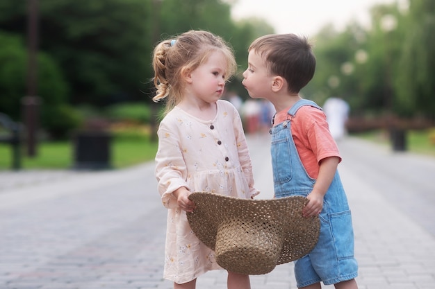 petite fille et garçon s'embrassent et s'embrassent ils se tiennent au centre de l'allée verte et tiennent un chapeau de paille