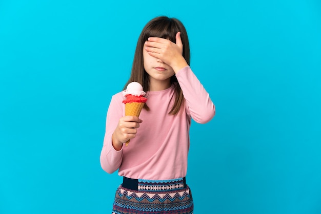 Petite fille avec une glace au cornet isolée sur fond bleu couvrant les yeux à la main. Je ne veux pas voir quelque chose