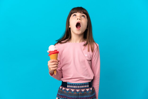 Petite fille avec une glace cornet isolée sur fond bleu en levant et avec une expression de surprise