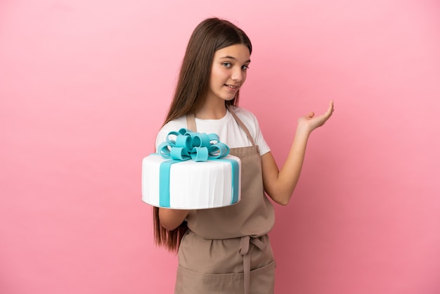 Petite fille avec un gros gâteau sur une surface rose isolée étendant les mains sur le côté pour inviter à venir