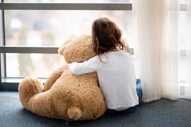 Une petite fille avec un gros ours en peluche est assise par terre et regarde par la fenêtre. Elle est bouleversée.