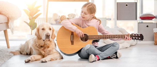 Petite fille avec guitare assise sur le sol dans une pièce ensoleillée avec chien golden retriever