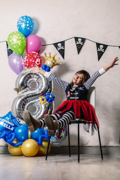 Petite fille habillée en pirate, les mains en l'air, assise sur une chaise, avec des ballons de célébration numéro huit