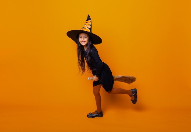 Petite fille habillée en sorcière d'Halloween dans une robe noire et un chapeau vole sur un manche à balai sur fond jaune.