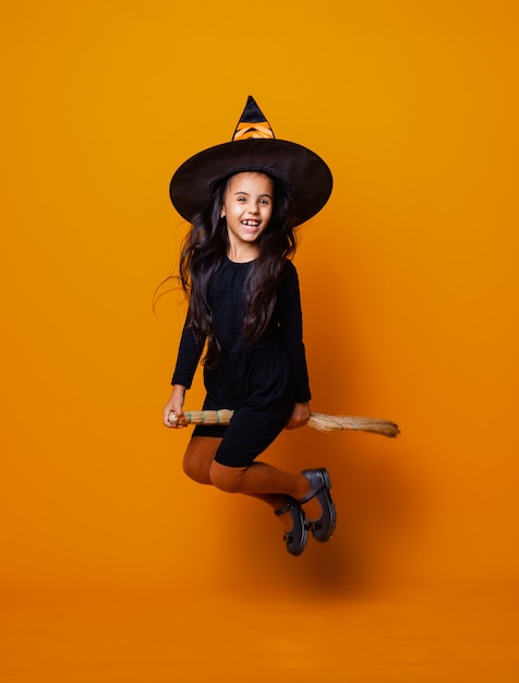 Petite fille habillée en sorcière d'Halloween dans une robe noire et un chapeau vole sur un manche à balai sur fond jaune.