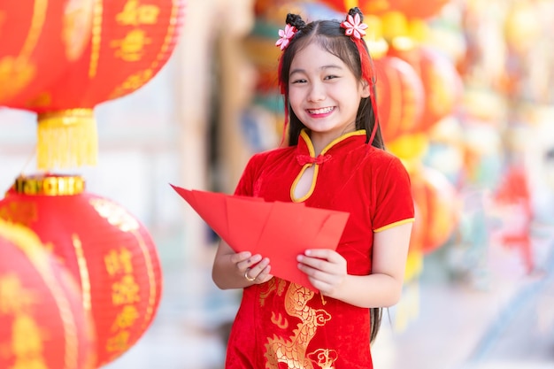 Petite fille heureuse asiatique portant une décoration cheongsam traditionnelle chinoise rouge tenant des enveloppes rouges à la main et des lanternes avec le texte chinois Bénédictions écrites dessus Est une bénédiction de fortune pour les chinois