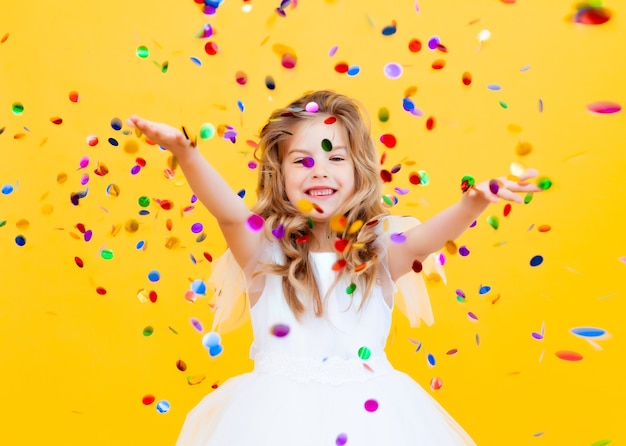 Une petite fille heureuse aux cheveux blonds et vêtue d'une robe blanche attrape des confettis sur fond jaune, concept de vacances