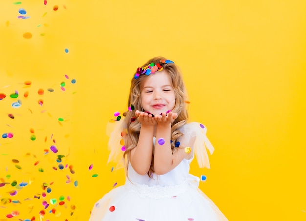 Une petite fille heureuse aux cheveux blonds et vêtue d'une robe blanche attrape des confettis sur fond jaune, concept de vacances