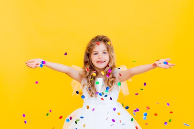 Une petite fille heureuse aux cheveux blonds et vêtue d'une robe blanche attrape des confettis sur fond jaune, concept de vacances