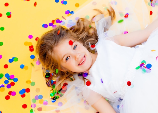 Une petite fille heureuse aux cheveux blonds et vêtue d'une robe blanche attrape des confettis sur fond jaune, concept de vacances