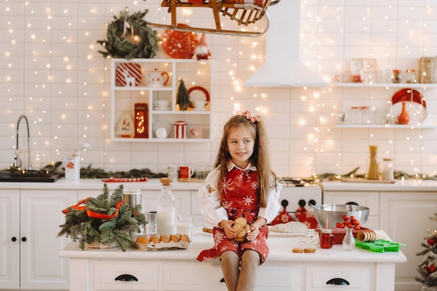 Une petite fille heureuse dans la cuisine de Noël est assise sur la table avec des biscuits dans ses mains.