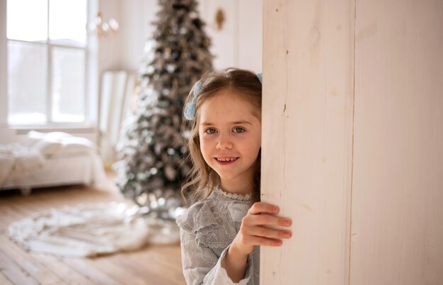 Une petite fille heureuse dans une robe en dentelle bleue se cache derrière une porte blanche dans une pièce