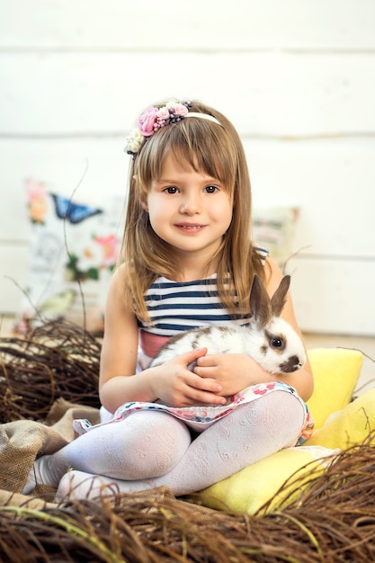 Une petite fille heureuse dans une robe est assise dans un nid et tient un joli lapin de Pâques blanc moelleux.