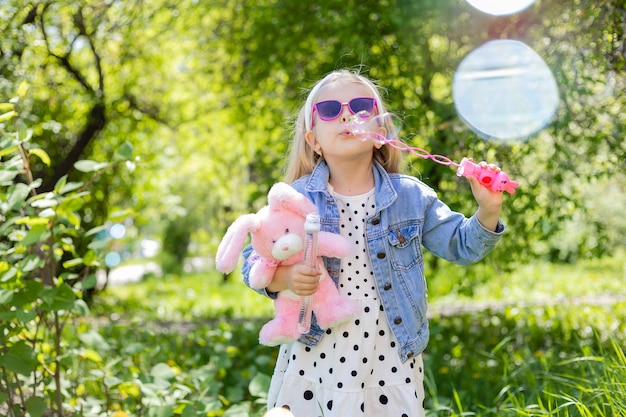 Une petite fille heureuse en été avec des lunettes de soleil gonfle des bulles de savon tient un jouet dans ses mains