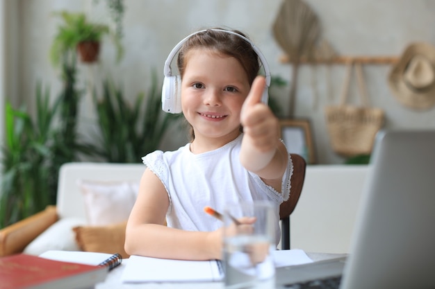 Une petite fille heureuse étudie en ligne à l'aide d'un ordinateur portable à la maison, un petit enfant souriant montre le pouce vers le haut, recommande une classe ou une leçon.