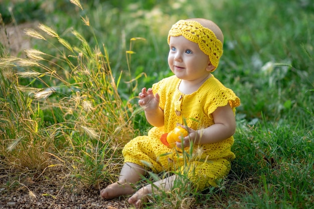 Petite fille heureuse mignonne assise sur l'herbe verte par une journée d'été ensoleillée vêtue d'une combinaison jaune en coton tricoté