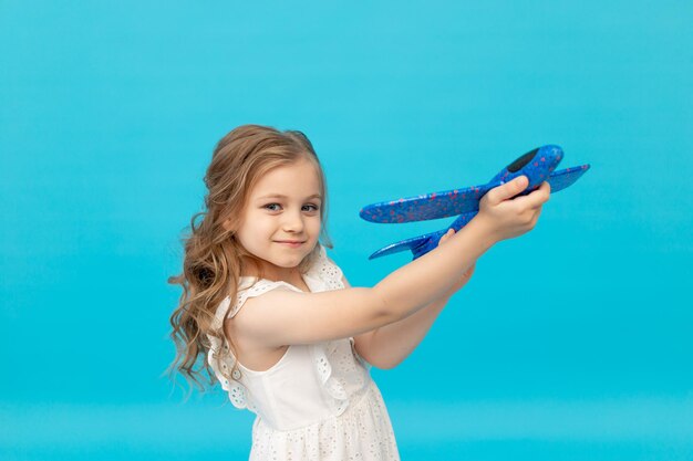 Petite fille heureuse mignonne dans une robe blanche de coton sur un fond bleu avec un avion dans ses mains jouant, un endroit pour le texte