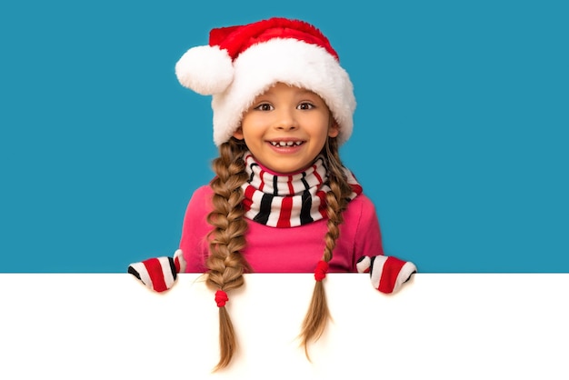 Une petite fille heureuse regarde derrière un mur blanc dans un chapeau de Noël.