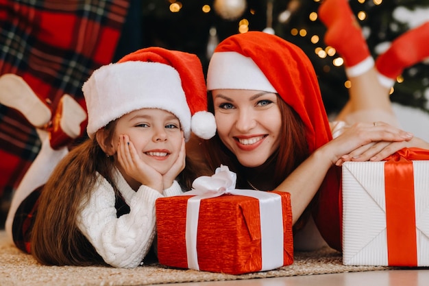 Une petite fille heureuse et sa maman en chapeaux de père Noël sourient avec des cadeaux à la main.