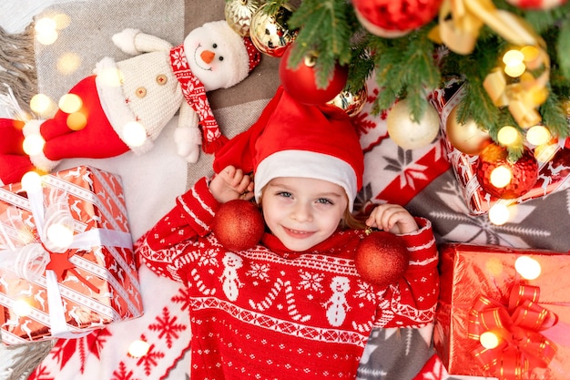 Une petite fille heureuse se trouve sous le sapin de Noël à la maison avec des boules de Noël et attend le nouvel an ou Noël dans un chapeau de père Noël rouge et sourit et se réjouit