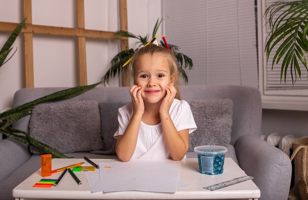 Une petite fille heureuse en T-shirt blanc est assise à une table avec des fournitures scolaires