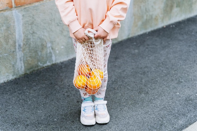 Une petite fille heureuse tenant des oranges dans un sac en maille près d'un mur de briques.