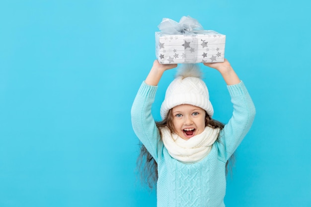 Une Petite Fille Heureuse En Vêtements D'hiver, Un Chapeau Et Un Pull Sur Fond Bleu Isolé Détient Des Cadeaux Pour Le Nouvel An Ou Noël Et Des Sourires, Un Endroit Ou Un Espace Pour Le Texte