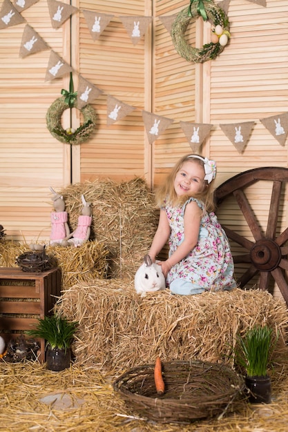 Une petite fille heureuse vêtue d'une robe est assise dans un nid et tient un joli lapin de Pâques blanc moelleux