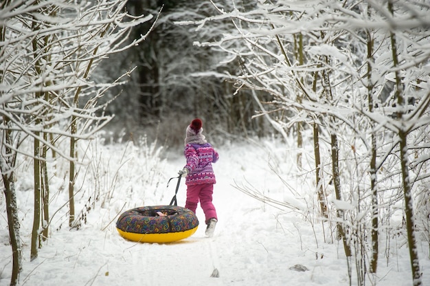 Une petite fille en hiver vêtue de vêtements violets et d'un cercle gonflable marche dans la rue dans une forêt enneigée.