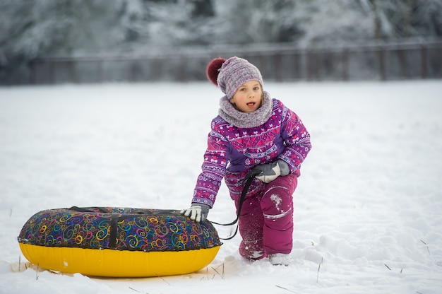 Une petite fille en hiver vêtue de vêtements violets et d'un cercle gonflable marche dans la rue dans une forêt enneigée.