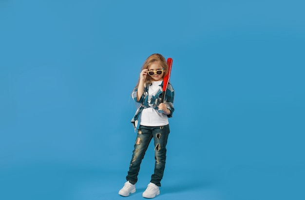 Une petite fille hooligan en jeans déchirés avec une batte de baseball dans ses mains sur un fond bleu isolé.