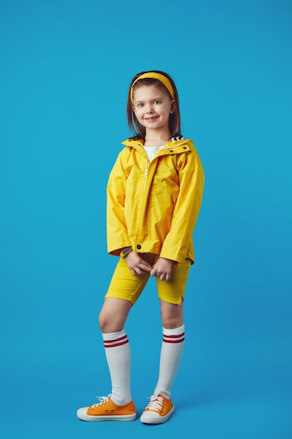 Petite fille en imperméable jaune et baskets sourit et garde les mains ensemble