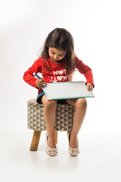 Petite fille indienne écrit sur ardoise avec marqueur, alors qu'il était assis sur un tabouret sur fond blanc