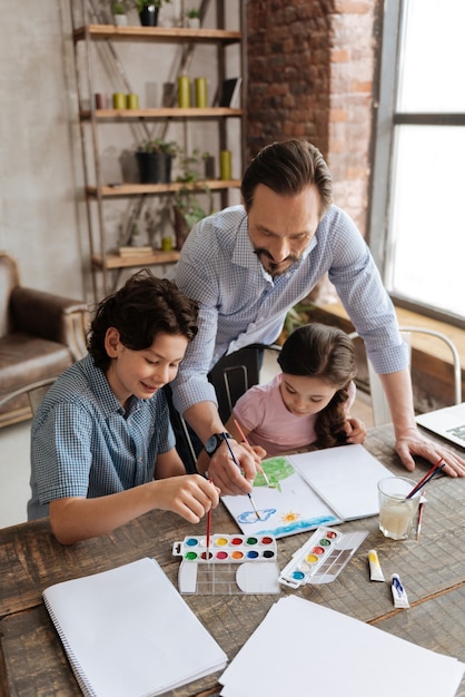 Petite fille inspirée et son père appliquant des coups de pinceau sur une peinture lumineuse pendant que son frère choisit la couleur suivante