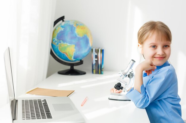 Petite fille intelligente fait ses devoirs dans son salon. Elle est assise à son bureau, écrit avec un stylo dans ses manuels et utilise un ordinateur portable.