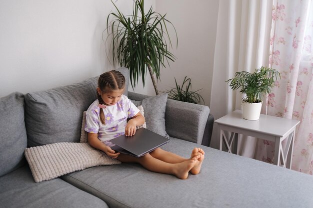 Une petite fille intelligente ouvre l'ordinateur portable de son père alors qu'elle est assise sur le canapé à la maison.