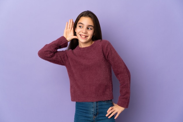 Petite fille isolée sur fond violet écoutant quelque chose en mettant la main sur l'oreille