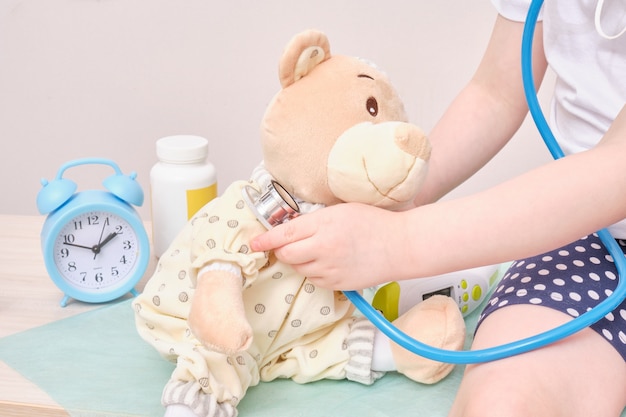 Petite fille jouant au docteur et à l'écoute de l'ours en peluche avec réveil stéthoscope et pilules sur fond