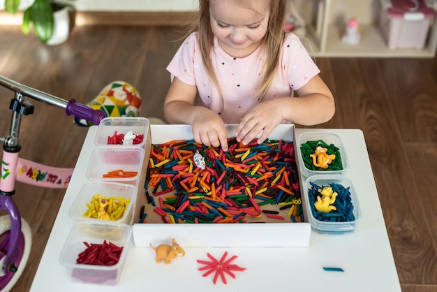 Une petite fille jouant avec un bac sensoriel avec des pâtes colorées pour des activités ludiques et manuelles Activité de jeu sensoriel et d'apprentissage des couleurs pour les activités des enfants Jeux Montessori pour la motricité fine