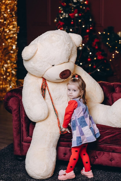 Une petite fille joue avec un gros ours en peluche sur le fond d'un sapin de Noël